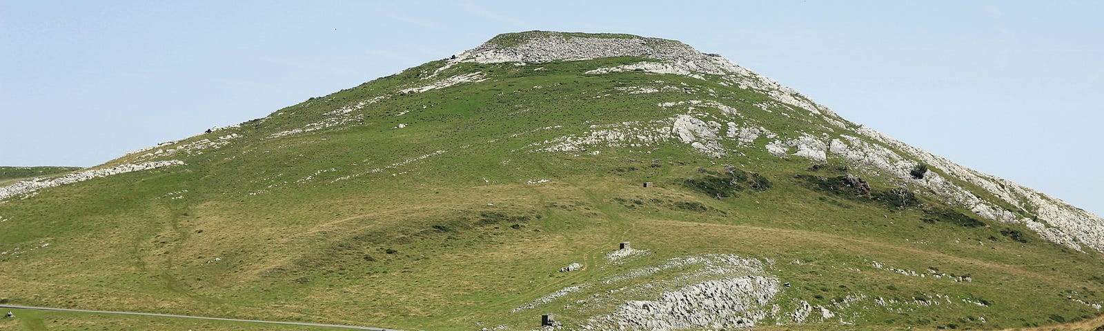 A flat-topped green, treeless, and rocky hill dominates the scene. A bit of pale blue sky. A narrow grey path bisects the scene, and a distant, lone walker heads toward the viewer.