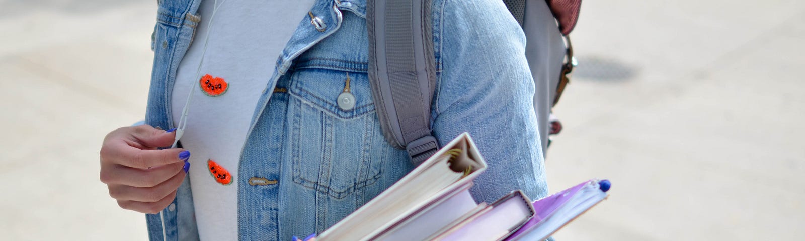 A student wearing a backpack, carrying textbooks in one hand and holding headphones in the other.