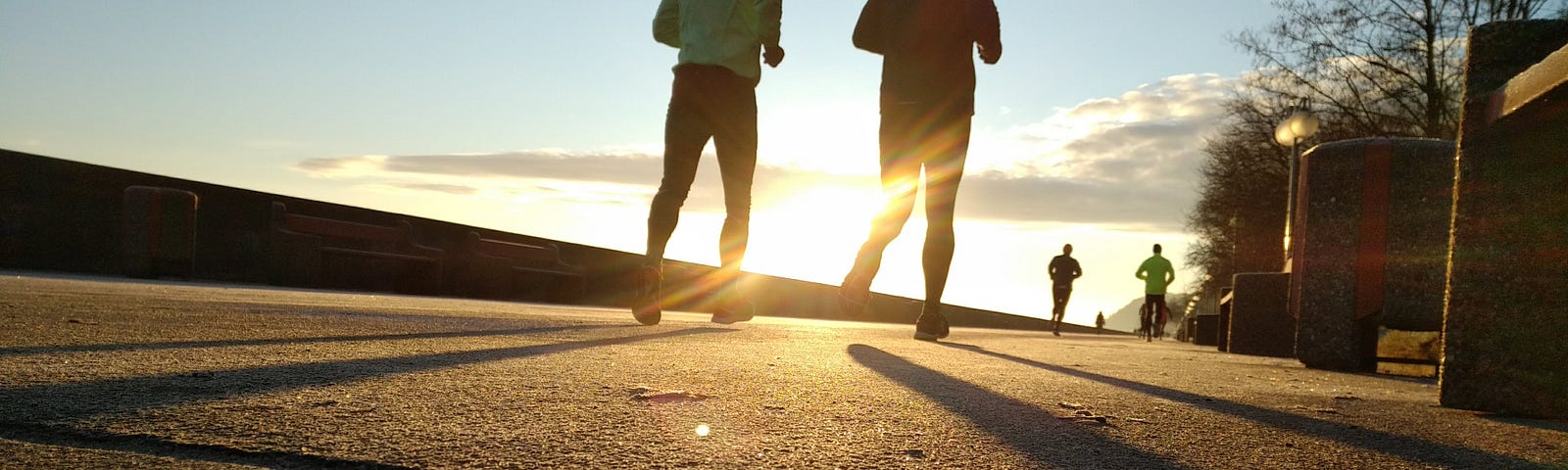 Silhouette of two runners wearing hats and gloves run away from the camera down a sidewalk at sunrise.