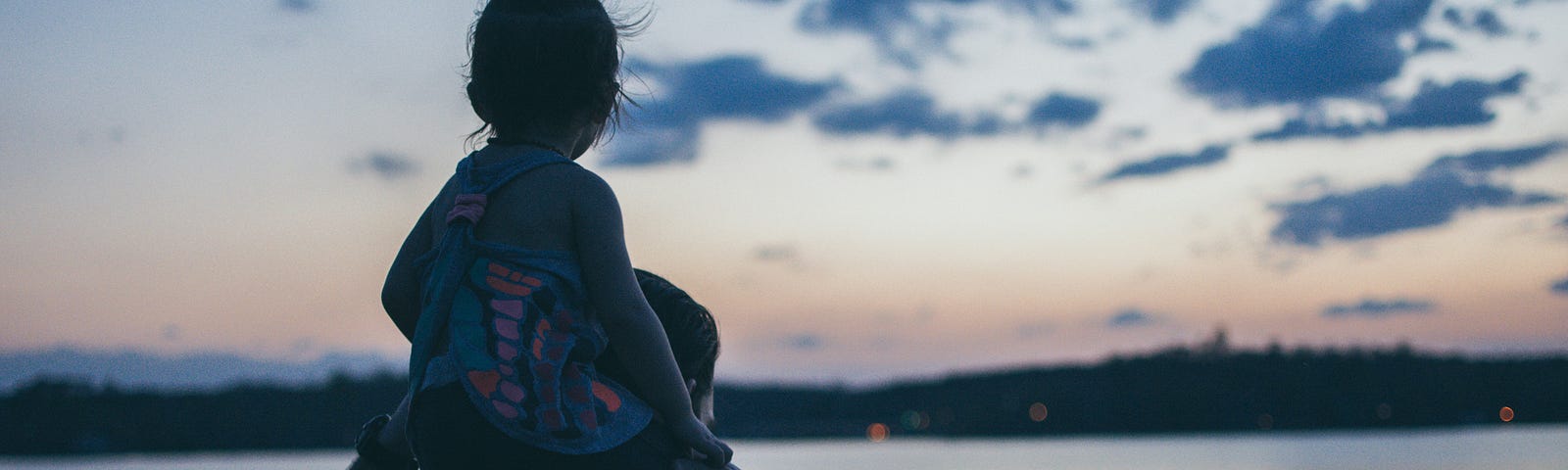 A sunset behind mountains and a lake. A father is holding his daughter on his shoulders and both of them are watching the nature in front of them.
