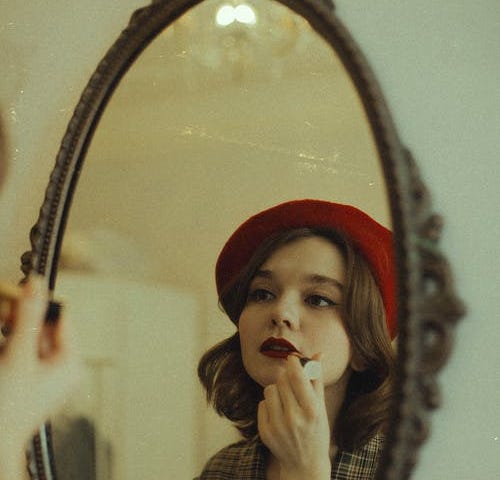 A woman, who is wearing a red beret, is applying red lipstick using a mirror.