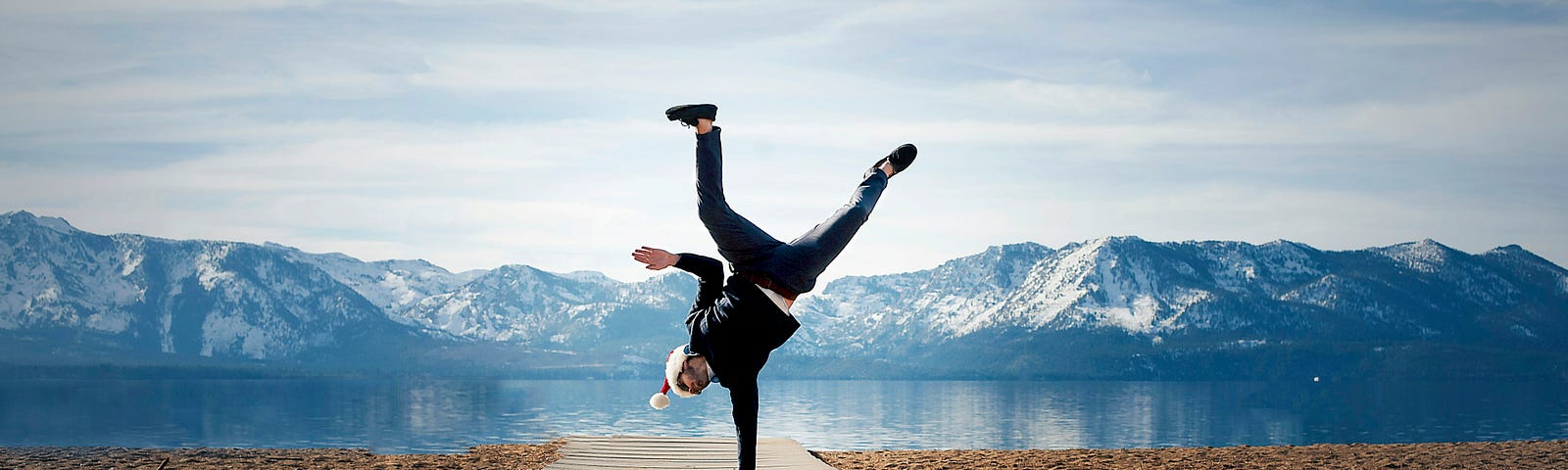 Man with Santa hat standing on one hand with a lake and mountains in behind him.