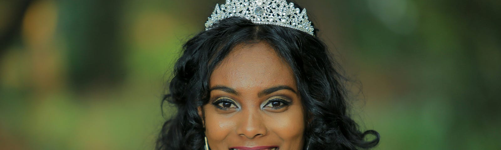 A smiling, beautiful young woman with long dark hair, wearing a tiara, and holding up red and white roses, as if at a prom.