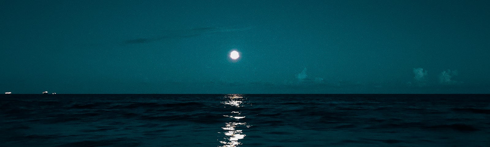 Photo of a beach at night, under a full moon. The sky is shades of dark turquiose, and the water looks darker with the night. A gentle wave meets the sandy beach with white motion in the dark night.