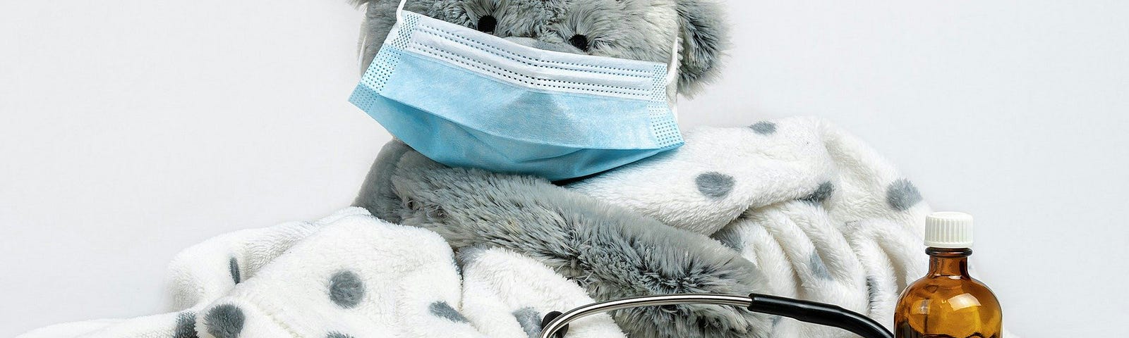 A gray teddy bear covered in a blanket and a face mask, with pills, a stethoscope, and cough syrup in the foreground