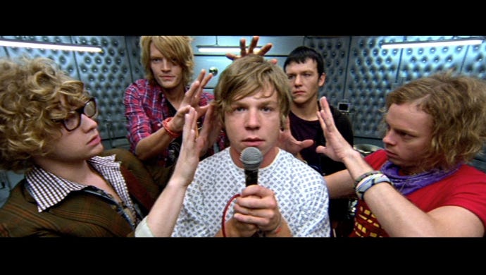 From “In One Ear” music video; Cage the Elephant band members surrounding frontman Matt Schultz with splayed hands around his head — Schultz staring at the camera wearing a hospital gown and holding a microphone with a padded room backdrop