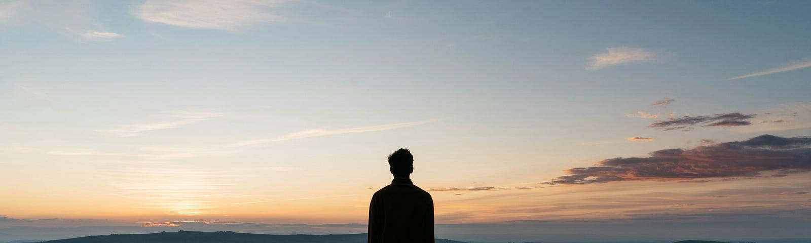 A person stands in silhouette against a vast sky painted with the subtle hues of a sunset, overlooking a tranquil landscape.