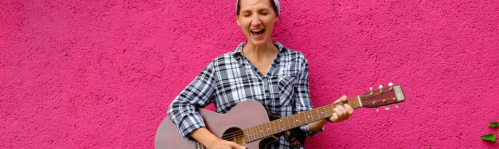 Woman standing against a pink wall and playing the guitar.