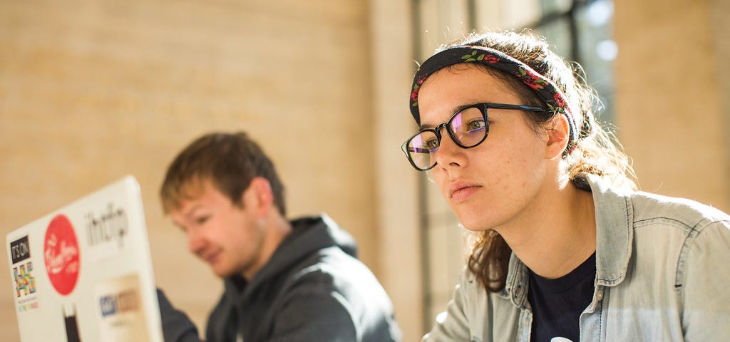A young woman wearing glasses works at a laptop, with a young man in the background next to her
