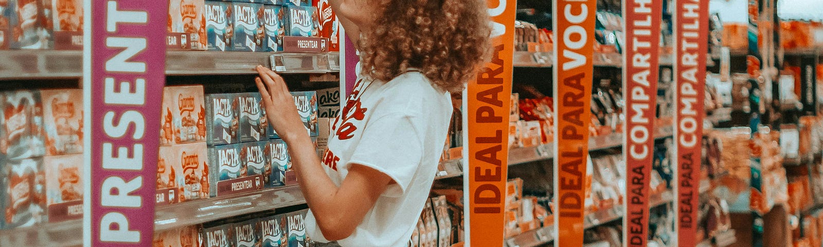 A lady with curly hair reaches to a high shelf to choose her product.