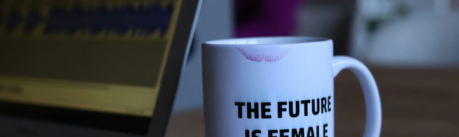 Photo of a white coffee mug that simply says “The Future Is Female” sitting on a table next to an open laptop.