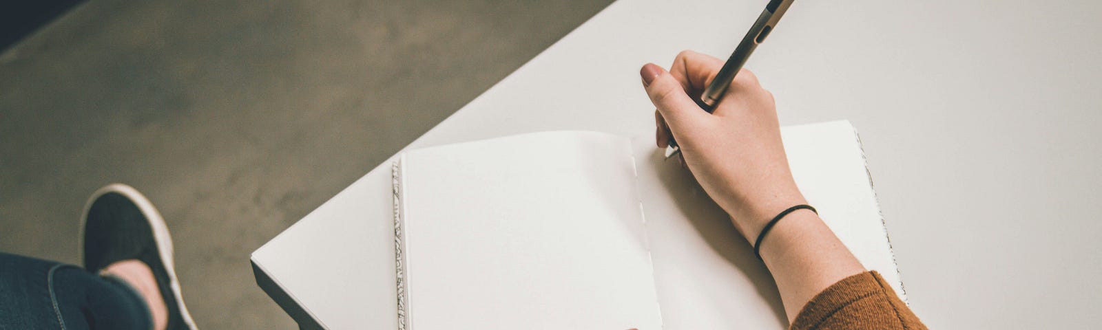 Person sitting on the left side of a white table with legs crossed wearing dark blue jeans and black shoes. The notebook is open with empty pages. The person has a pen in hand ready to write.