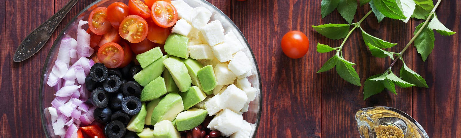 A plate with vegetables, on a dark wood table, with a fork, three grape tomatoes, and some green mint leaves.