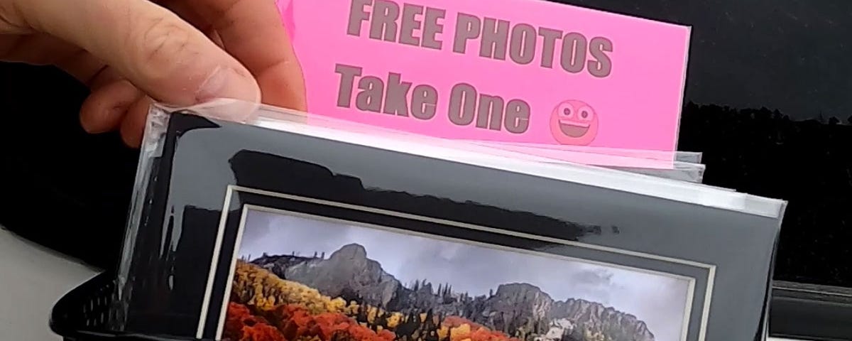 A hand reaching into a bin filled with 5x7 matted photos with a sign that says “Free Photos — Take One”