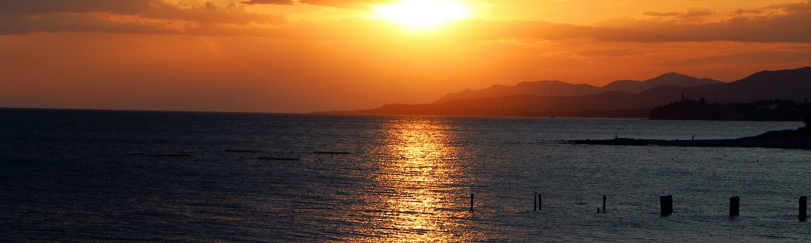 The light of the orange setting sun is reflecting on the sea water and the beach. In the background, dark silhouettes of mountains are visible.