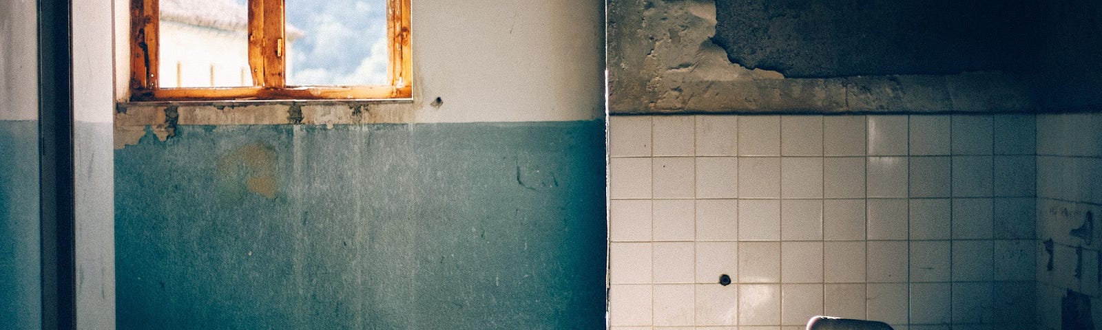 A partially daylit bathroom showing the remains — broken tiles, fallen sink on the floor, a broken window, and peeling paint.