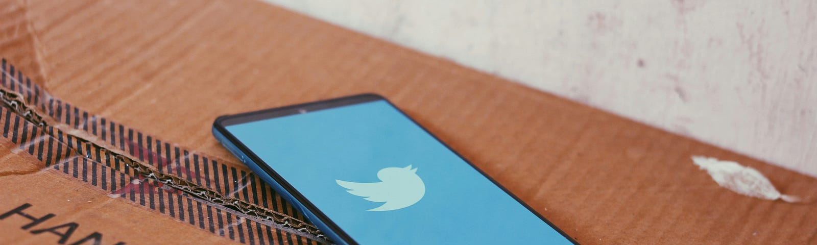 A phone with the twitter bird logo on its screen sits on a cardboard box with the words “handle with care” on it.