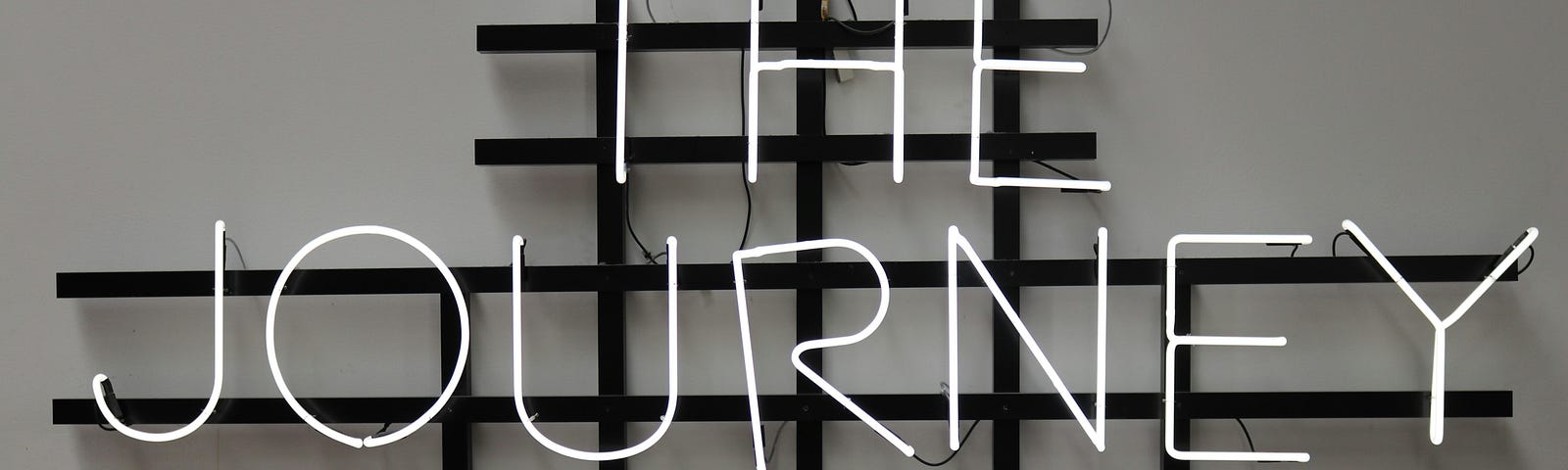 A black and white photograph of a neon sign that says “The Journey is On” in capital letters.