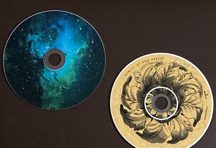 Two CDs on dark brown background; Panic! at the Disco “Vices and Virtues” on bottom right (gray outline of leaf flower on yellowed-white background and band name on portion of edge of the disc), CD at top left with blue/green nebula design