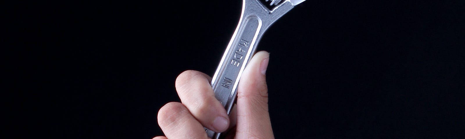 Man’s hand holding up a wrench, against a black background.