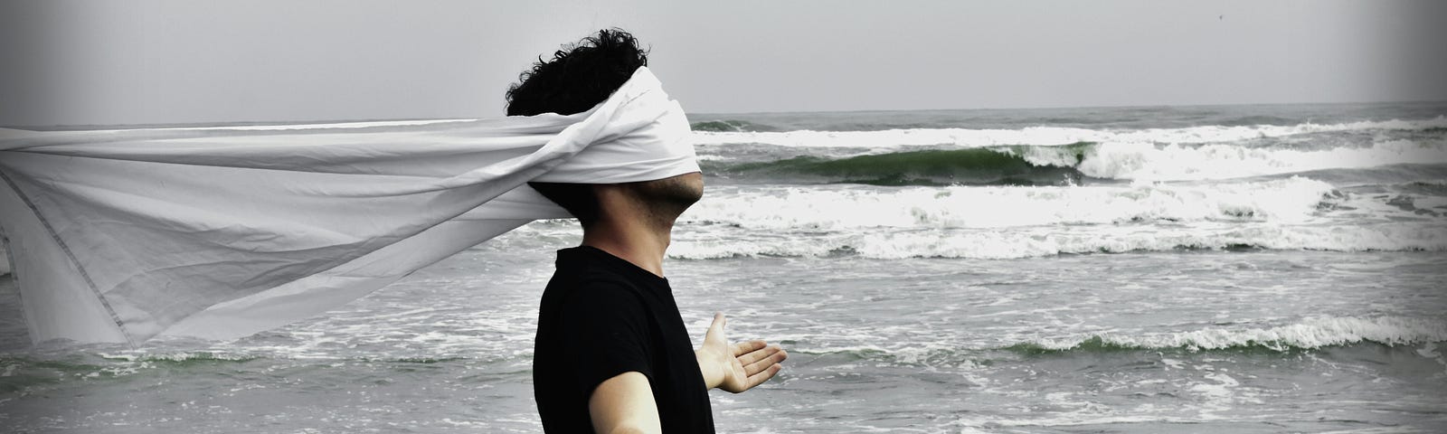 man blinfolded with a white scarf in front of the ocean.
