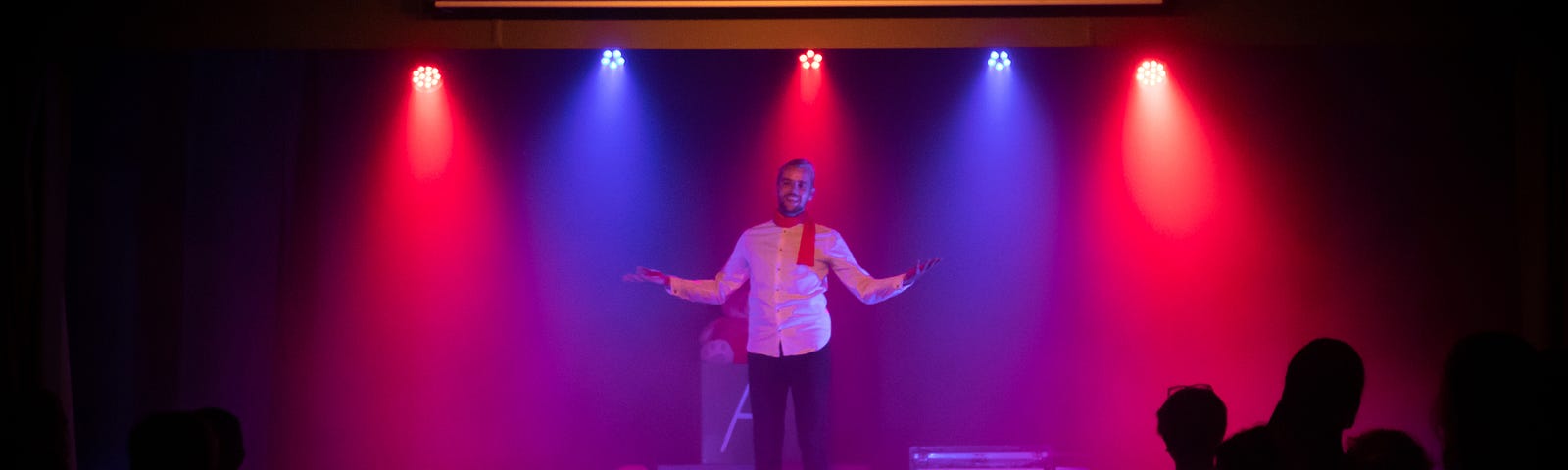 A white man with a beard wearing a white collared shirt, red scarf, and dark pants stands on a stage backlit by alternating blue and red lights in an otherwise dark room. He has his arms slightly outstretched. The dark silhouettes of a few people in the audience can be seen.