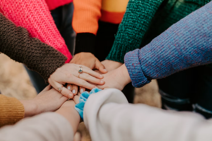 Several hands stacked on top of each other in a circle. The hand on top of the huddle has a ring on their thumb, middle, and ring finger. The sleeves of their sweaters are orange, green, blue, and white.