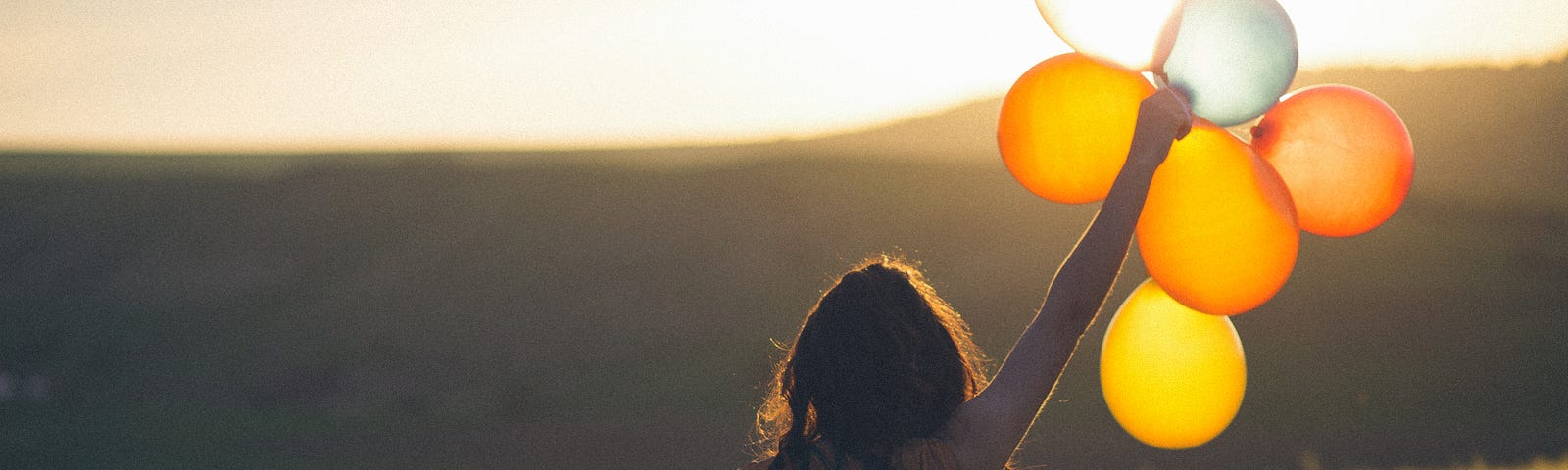 Person holding a bunch of balloons and looking into the sunset, on a summer day in the field.