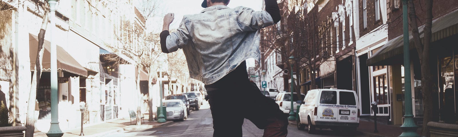 A young man grooves in the middle of a street, sunlight streaming down to touch the buildings and budding trees.