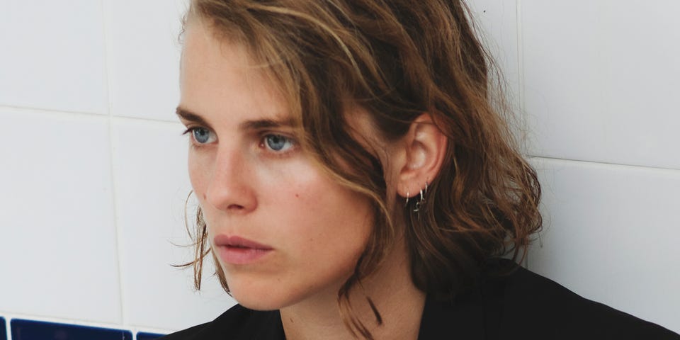 A photograph of English musician Marika Hackman. The photo shows her from the shoulders up. She is leaning against a tiled wall, with the lower tiles coloured dark blue and the upper tiles are coloured white. Marika Hackman has neck-length dirty blonde hair, pale skin, and dark eyebrows. She is wearing a black jacket and white shirt. She is facing to her right and looking off-camera with a neutral expression.