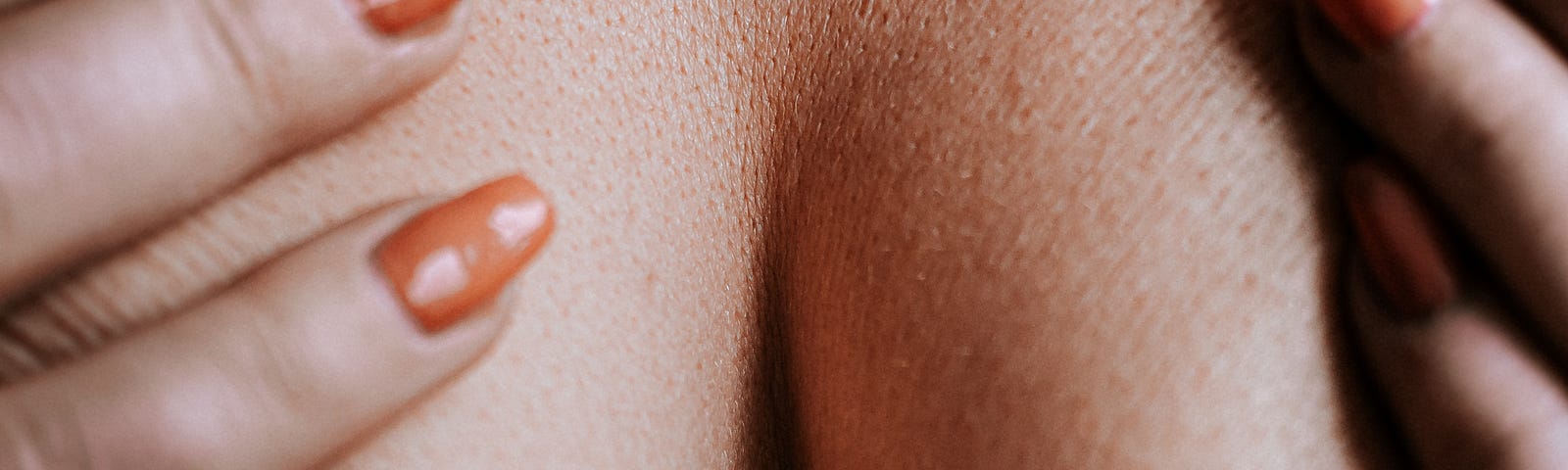 A close-up of the breast crack from a woman covering her bare ample breasts with orange nail-polished hands.