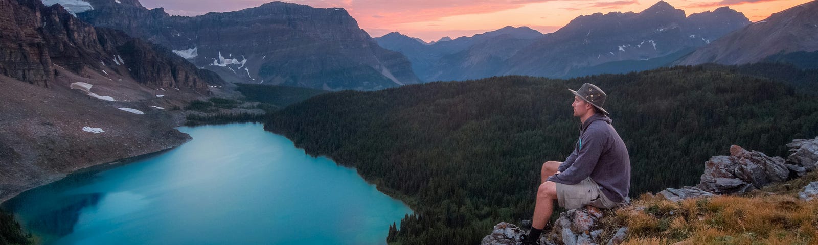 A man with a hat sitting alone surrounded by mountains, and a deep-blue lake in front of him