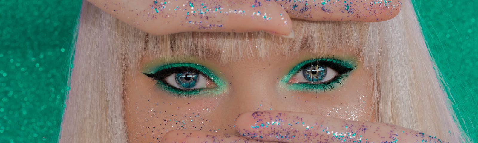 A blond with blue eyes and blue glitter-wiped palms facing out covering her nose and forehead to reveal only her heavily green-shadowed and eyelined eyes.