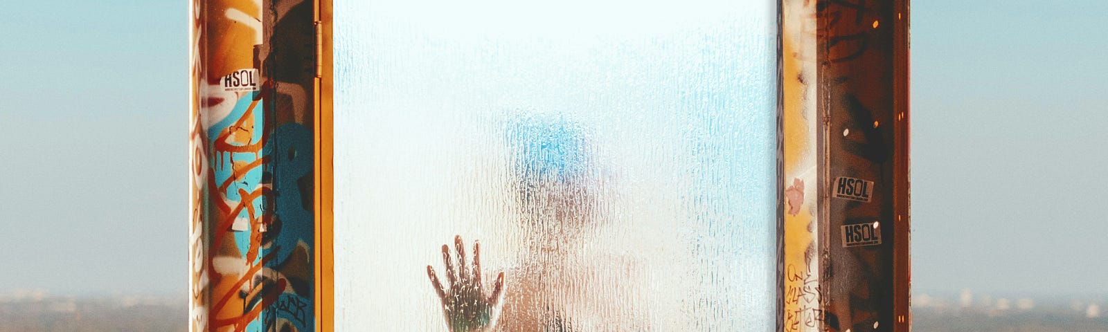 A woman, wearing only underwear, is encased in a frosted glass cube that is surrounded by graffiti. Her silhouette and garments are barely visible, but her palm, pressed against the glass, is clear to the observer. Her head is turned. away from us.