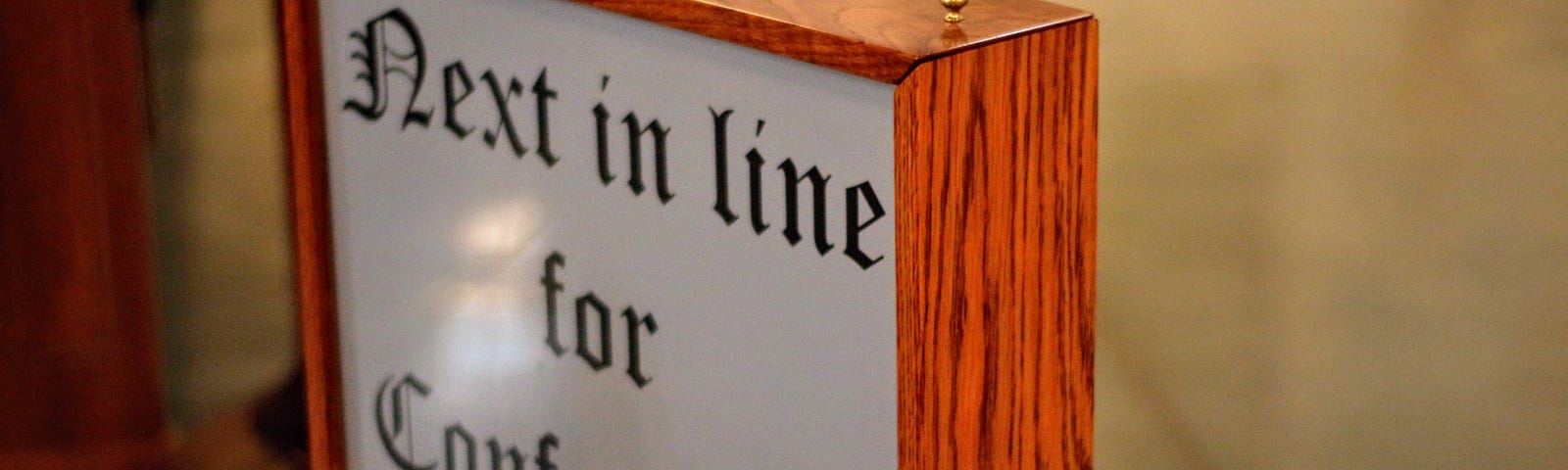 A white sign with a wooden border and antique lettering that reads, “Next in line for Confession”