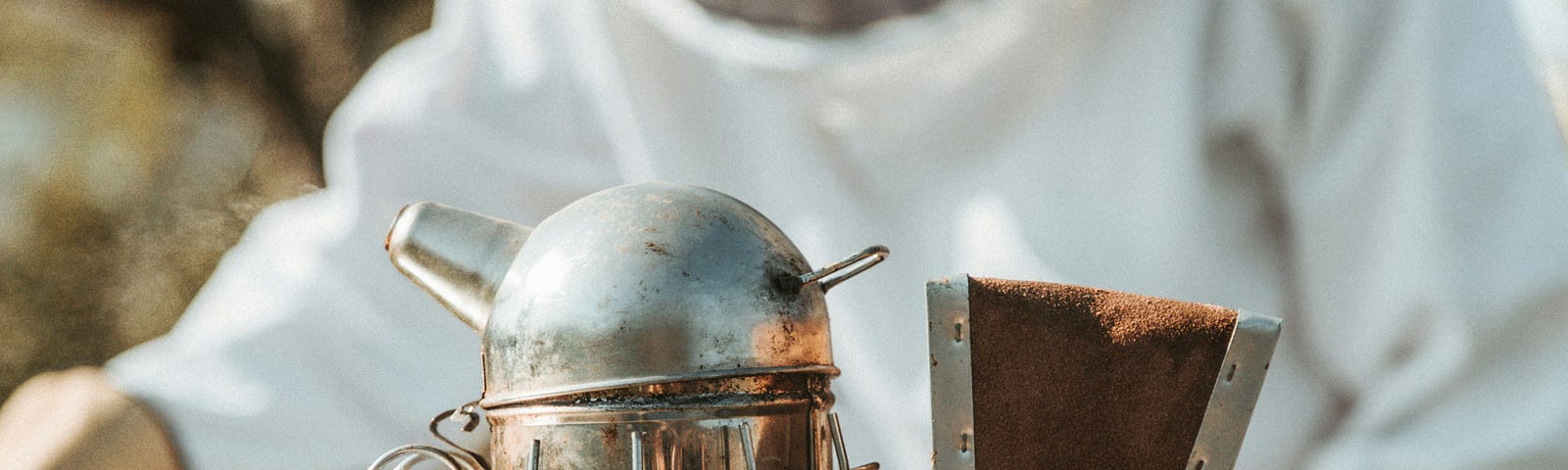 Photo of a beekeeper in white overalls and veil in the background, and a close-up of a smoker in the foreground.