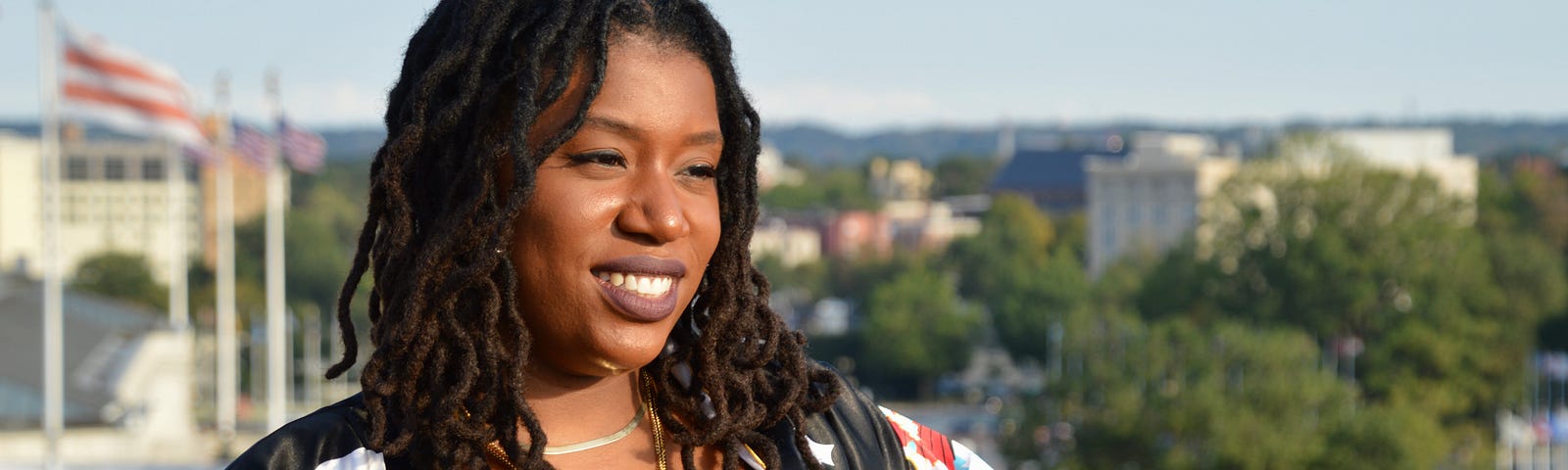 A smiling, mid-aged, African-American woman with shoulder length locks, posing outside during a bright, clear day.