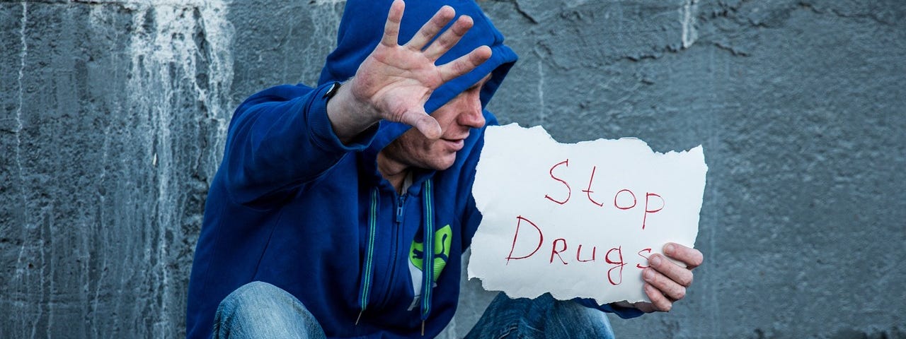 Young white man in a blue hoodie and jeans, sitting with knees up next to a gray concrete wall, holding his hand up with a sign that says, “Stop drugs”