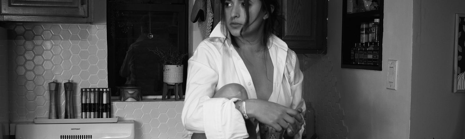 A black and white picture of a  woman seating on a chair in her kitchen, looking in the distance.