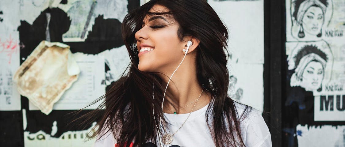 Woman with dark hair smiling and looking off to the side with a white t-shirt on, headphones in, and some designs and doodles pinned on a wall behind her