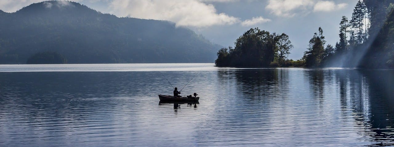 A cloudy day on the lake. A person is fishing in a small boat with trees and mountains in the background.