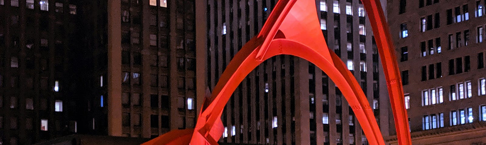 a nighttime photo of the huge bright-red sculpture in front of a tall office building