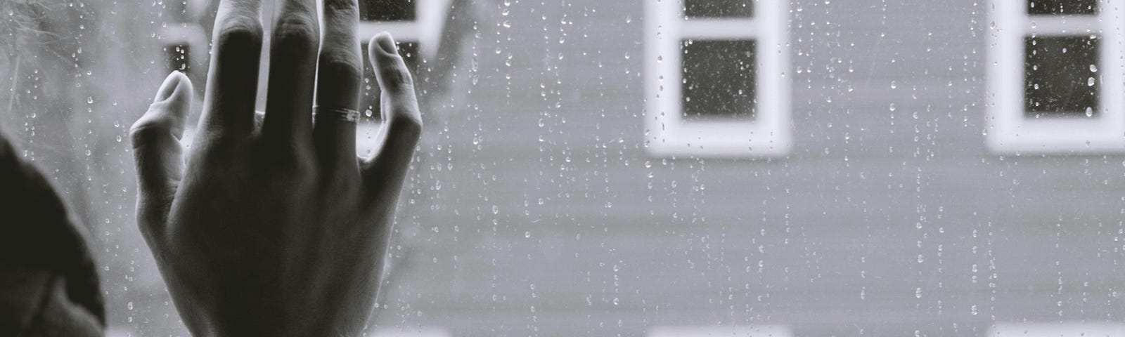 A person in front of a window, pressing their hand against the window pane as rain splatters on the glass.