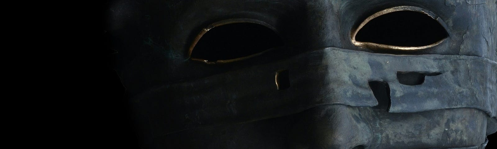 A grey, tattered, mask with gold around the eyes, on a black background