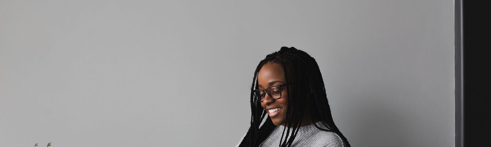 A black woman with braids is sitting and reading a black book with a white up in her hand. Next to her is a table with a plant on top.