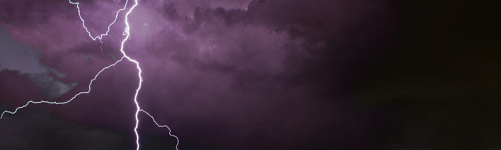 Lightening, evidenced by multiple white crooked lines, on a purple and dark shaded background of clouds.