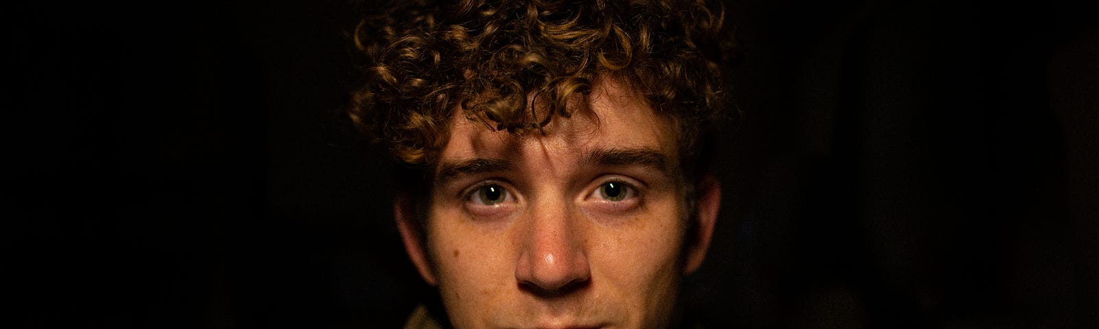 A boy with brown jacket and curly hair, standing in dark with slightly wet eyes.