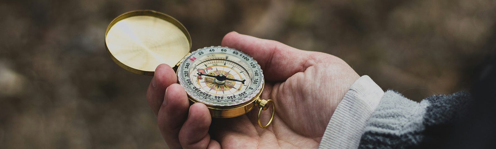 A person’s hand holds an old, gold compass, against a backdrop of autumn leaves and layered sleeves.