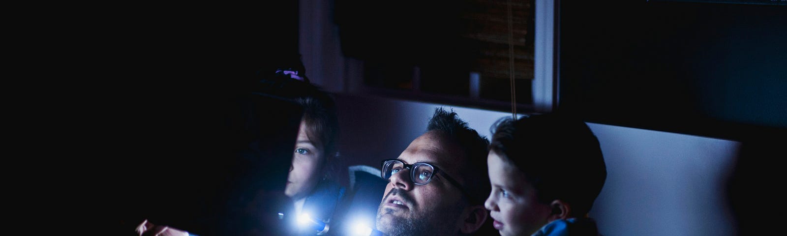 A father and his two young children are sitting in bed at night, illuminated by the light of a tablet or book they are looking at together. The father, wearing glasses, is in the middle, with a child on each side, all appearing engaged and absorbed in the story. The room is dark, with the soft glow of the screen lighting their faces, creating a cozy and intimate atmosphere.