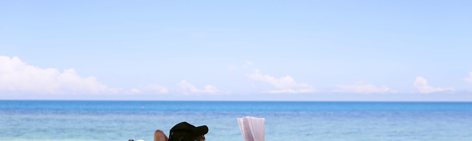 A woman in beach wear, sitting on a beach chair, reading a book, facing the sea.